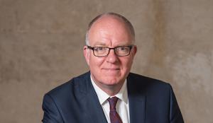 A man faces the camera. He is wearing glasses, a white shirt, red tie, and navy jacket. He is against a plain brown background.