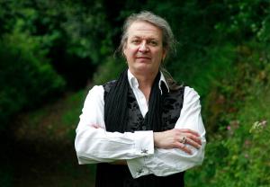 A man faces the camera. He is wearing a white shirt and a black waistcoat and scarf, standing outside in front of shrubbery. 