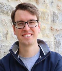 A man in glasses faces the viewer and smiles. He is wearing a navy blue jumper and pale blue t shirt.