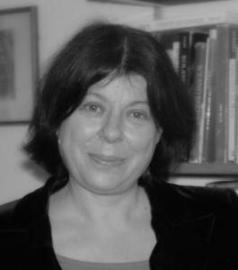 A black and white photo shows a headshot of a woman staring at the camera. She is wearing a black top and is stood in front of a bookshelf.