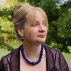 profile of blond-haired woman wearing a sheer black top and dark blue necklace. Trees in background