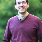 A man in a burgundy jumper smiles at the camera. He is against a blurred nature backdrop.